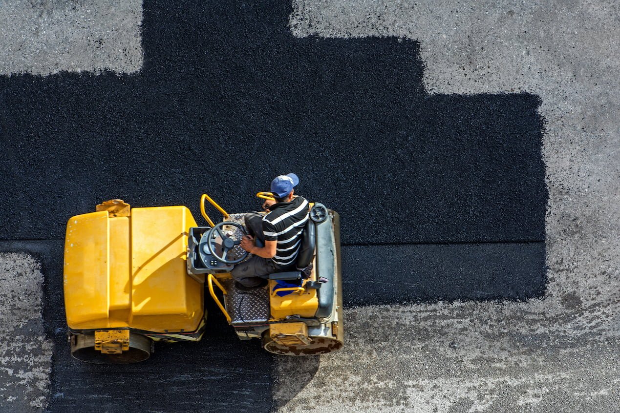 Armor Asphalt | Charlotte, NC | A worker in a striped shirt and blue cap operates a yellow paving machine, laying fresh black asphalt on a road. The image, taken from an overhead perspective, showcases the contrast between the new asphalt and the existing surface. This project is undertaken by Armor Asphalt In Charlotte, NC.