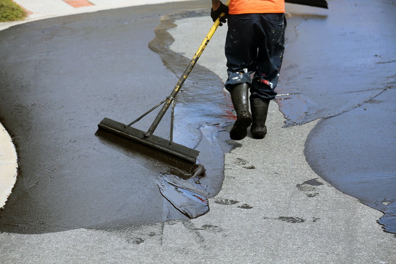 asphalt repair benefits Armor Asphalt | Charlotte, NC | A worker in an orange shirt and black boots, representing Armor Asphalt in Charlotte, spreads fresh asphalt on a curved NC road using a long-handled squeegee. The worker’s back is turned to the camera, and wet footprints are visible on the newly coated surface.