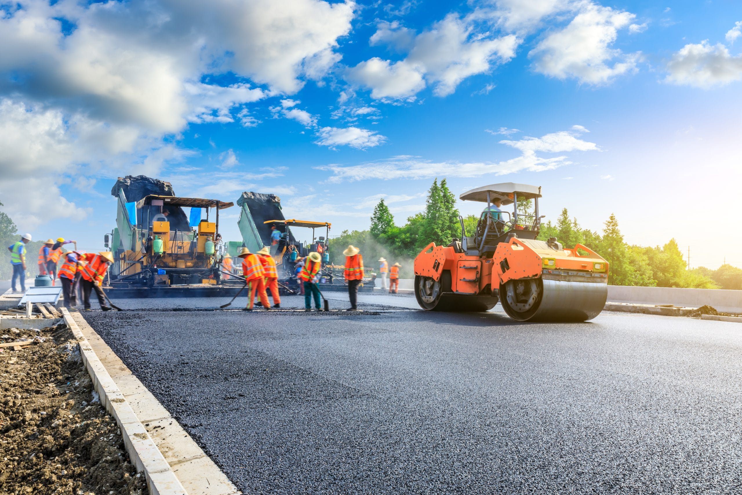 Construction workers in orange vests pave a road with machinery under a blue sky with clouds.