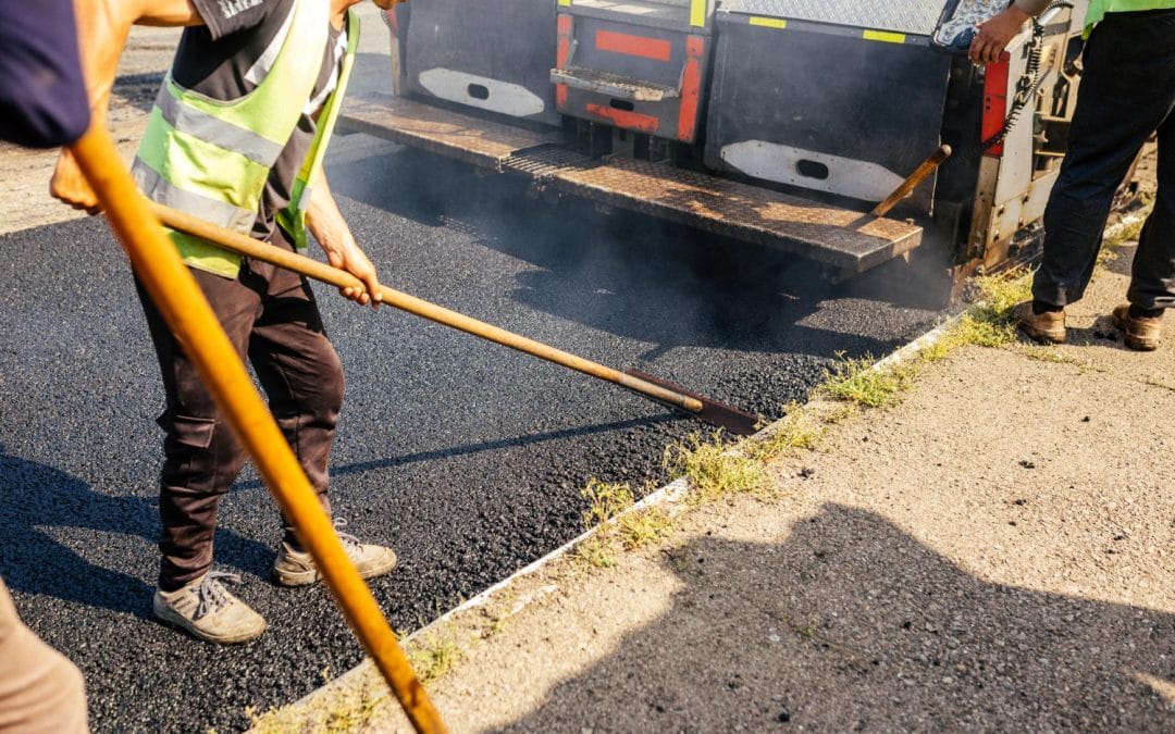 Workers use tools to spread hot asphalt on a road during paving, with a truck in the background.