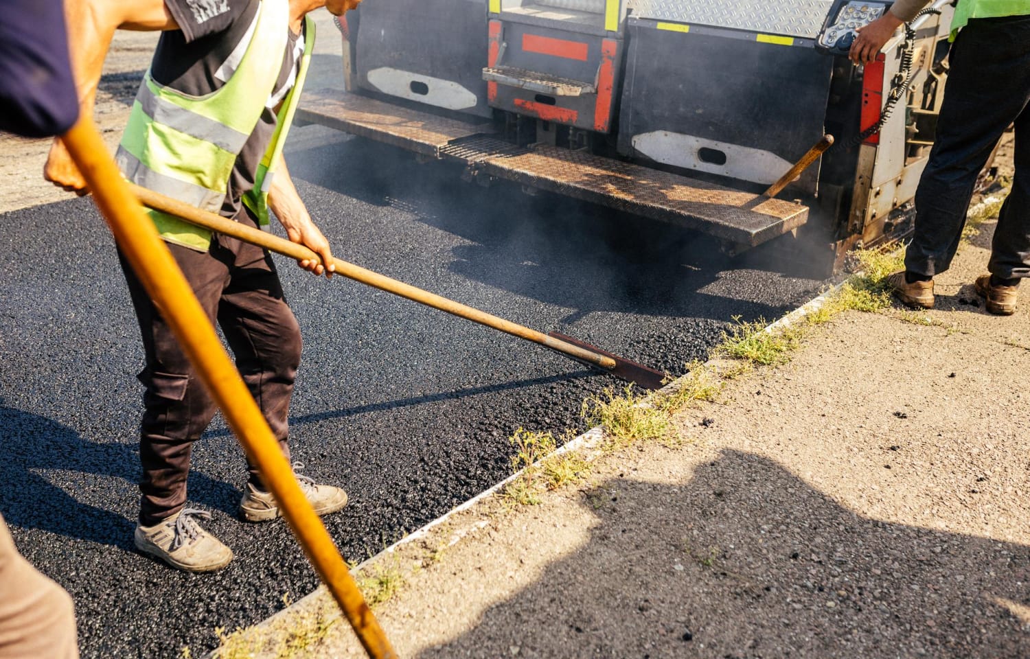 Workers use tools to spread hot asphalt on a road during paving, with a truck in the background.