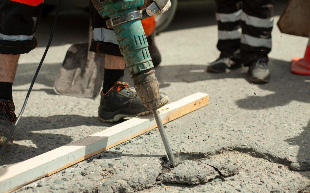 Worker in safety boots using a jackhammer to break asphalt on a road; legs and tools visible.