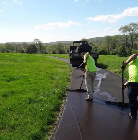 Two workers in safety vests spread fresh asphalt on a narrow road in a grassy rural area.